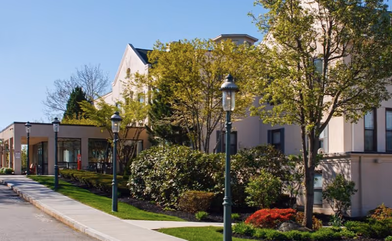 Exterior view of a senior living facility building with beige walls, multiple windows, and a well-maintained garden with trees, bushes, and flowers along a sidewalk with vintage-style street lamps.