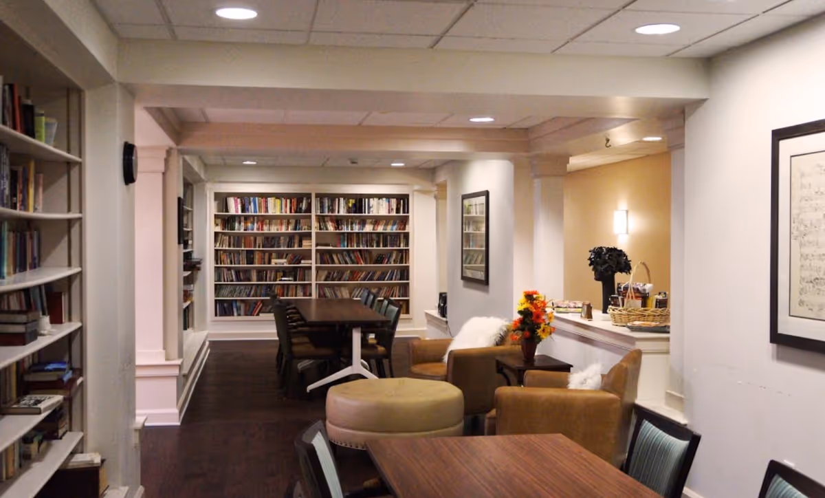 Interior view of a senior living facility common area featuring bookshelves filled with books, a long table with chairs, comfortable armchairs with cushions, a round ottoman, and a small table with a vase of flowers. The space is well-lit with ceiling lights and wall sconces, and has a warm, inviting atmosphere.