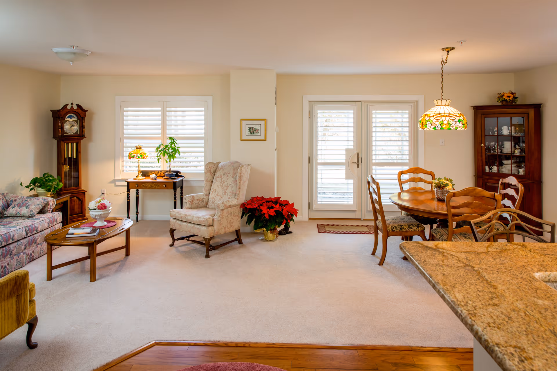 A cozy living and dining area with beige walls and carpet. The living room features a floral patterned sofa, a wooden coffee table with books, a floral armchair, a wooden grandfather clock, and a small table with a stained glass lamp and a plant. The dining area has a wooden round table with four chairs, a hanging stained glass lamp, and a wooden china cabinet filled with dishes. French doors with white shutters lead outside, and a granite countertop is visible in the foreground.