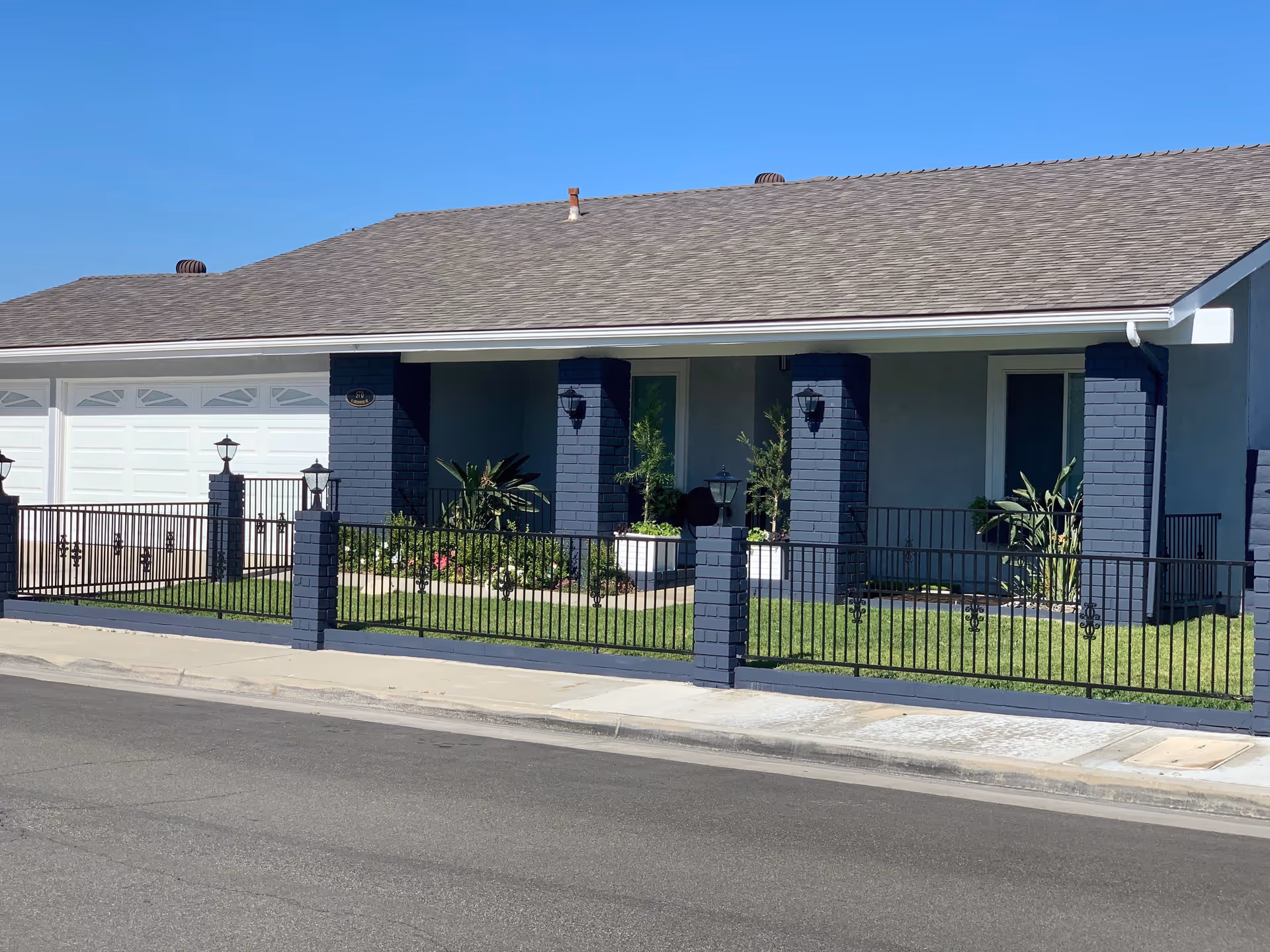 Front exterior view of a single-story building with a gray shingled roof, blue brick pillars, a black metal fence, and a well-maintained lawn with plants and flowers under a clear blue sky.