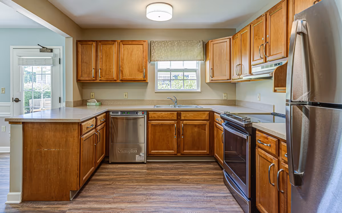 A clean and modern kitchen with wooden cabinets, a stainless steel refrigerator, dishwasher, and oven. There is a window above the sink with a patterned valance, and the floor is wood-style laminate. A door with glass panels leads outside.