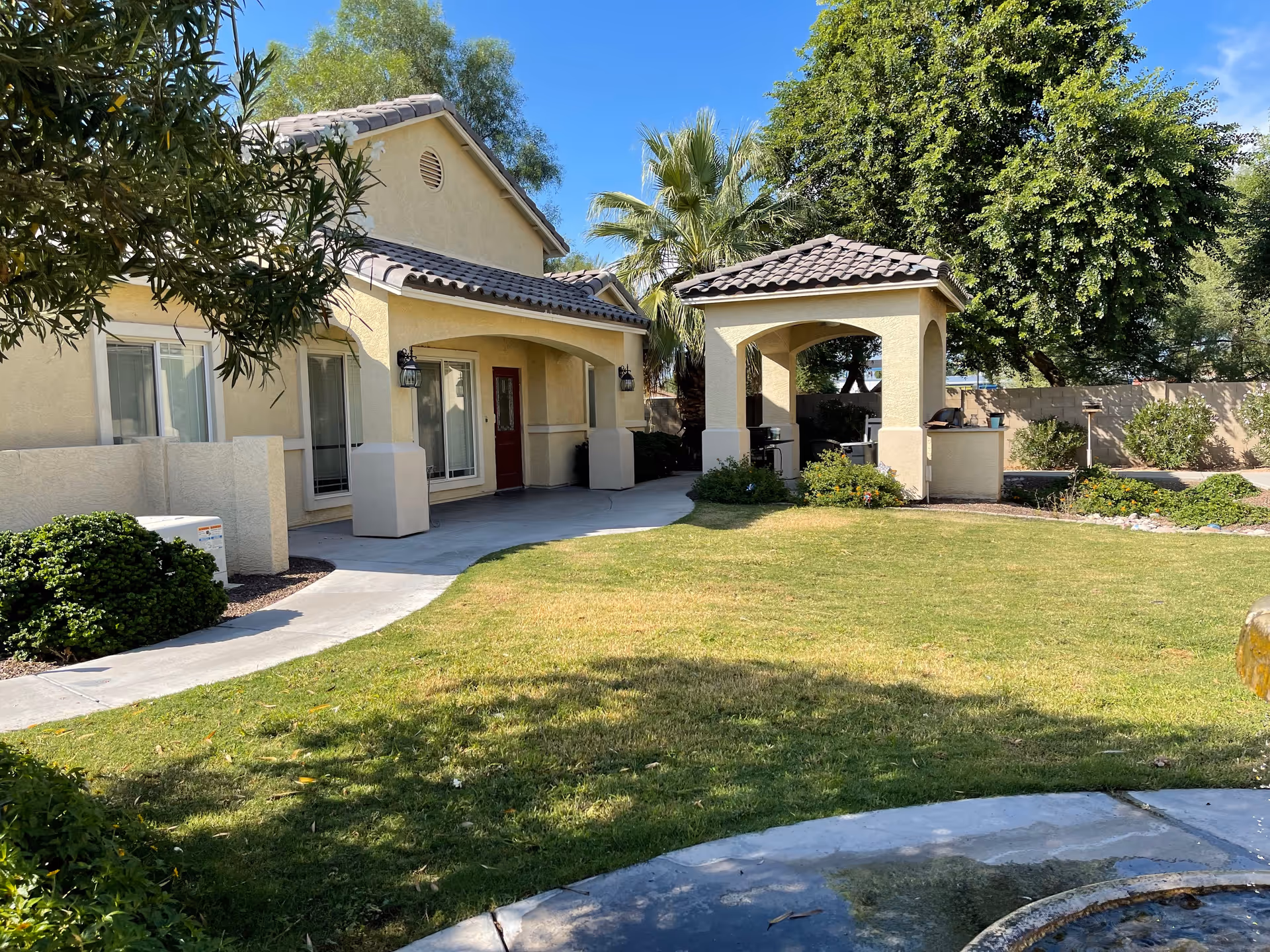 Beige assisted-living building with arched covered entryways, a small pavilion, and a grassy courtyard with trees.