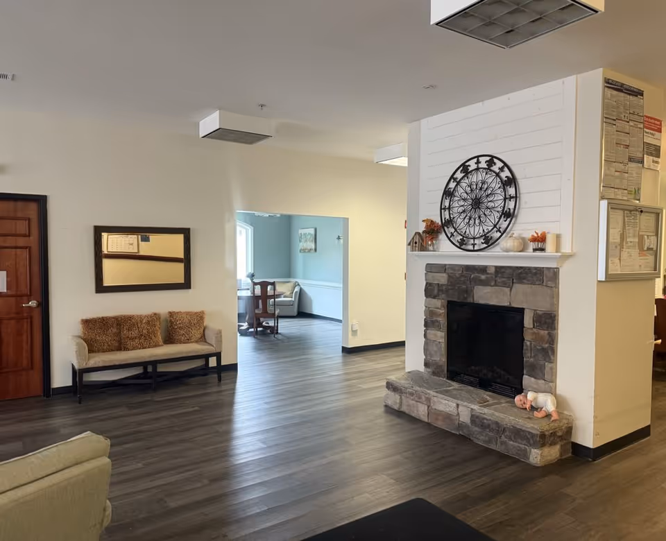 Interior view of a senior living facility common area with a stone fireplace decorated with a metal wall art, small pumpkins, and candles. There is a bench with two brown cushions against a wall with a mirror above it. The floor is wooden, and there is an open doorway leading to another room with a table and chairs.