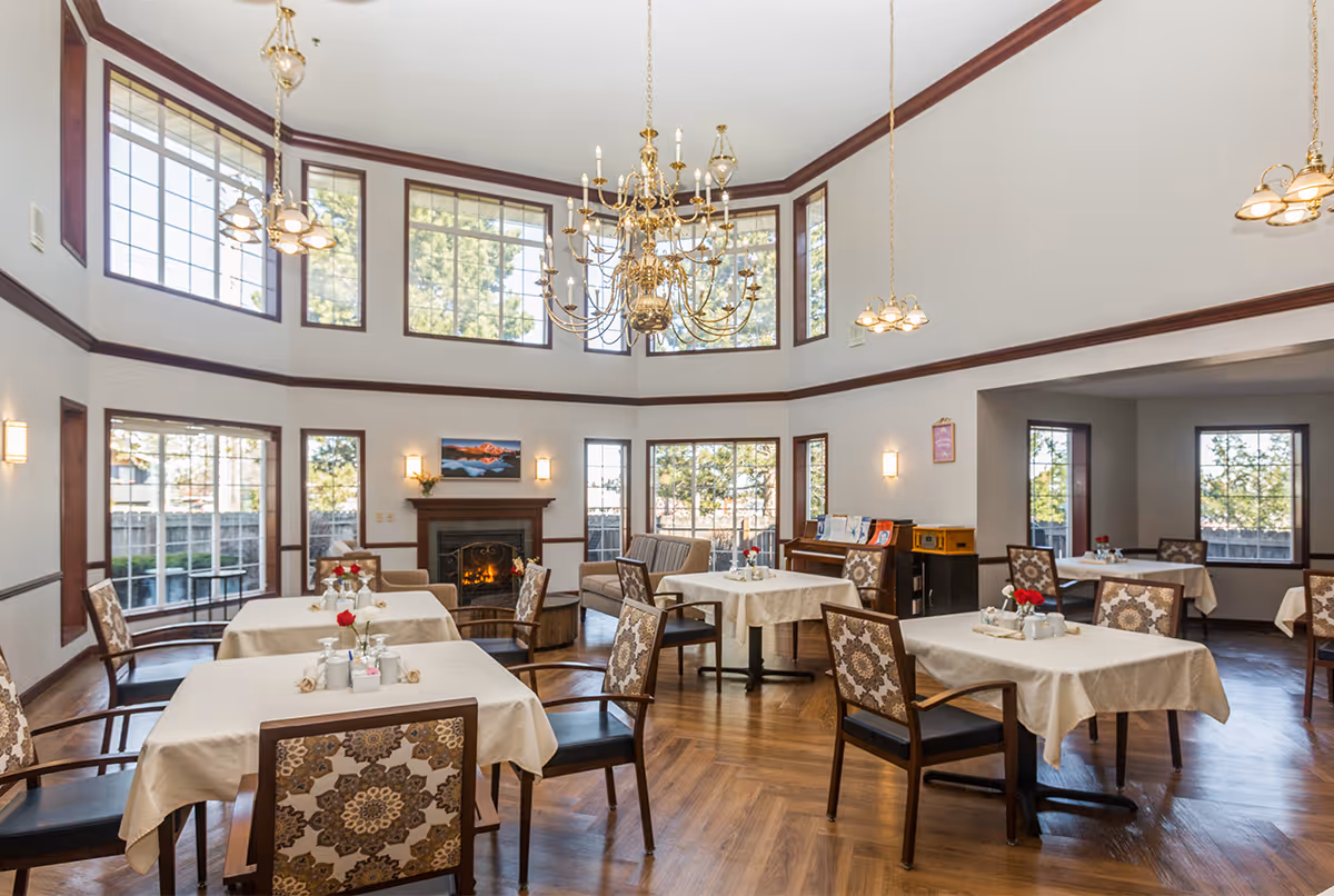 A bright and spacious dining room with multiple tables covered in beige tablecloths, each set with a small vase of red flowers and condiments. The room features large windows allowing natural light to fill the space, a fireplace with a painting above it, elegant chandeliers hanging from a high ceiling, and wooden flooring. There are patterned chairs around the tables and a piano against one wall.