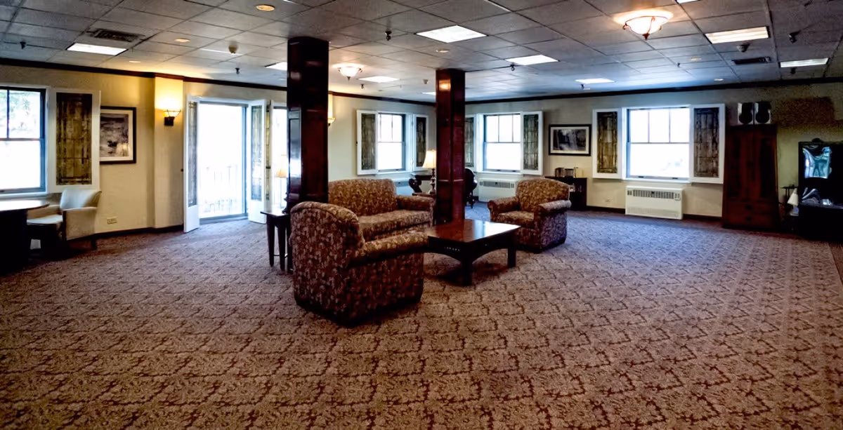 Spacious senior living common room with patterned carpet, upholstered sofas and armchairs arranged around a central coffee table and windows along the far wall.