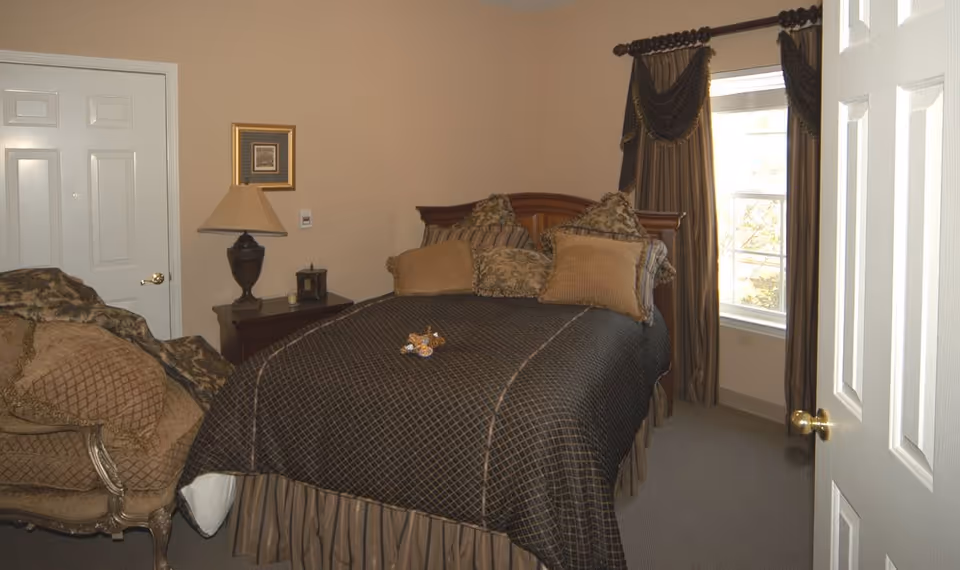 A neatly furnished bedroom with a bed covered in patterned bedding, multiple pillows, a bedside table with a lamp, an armchair, and a window with drapes.