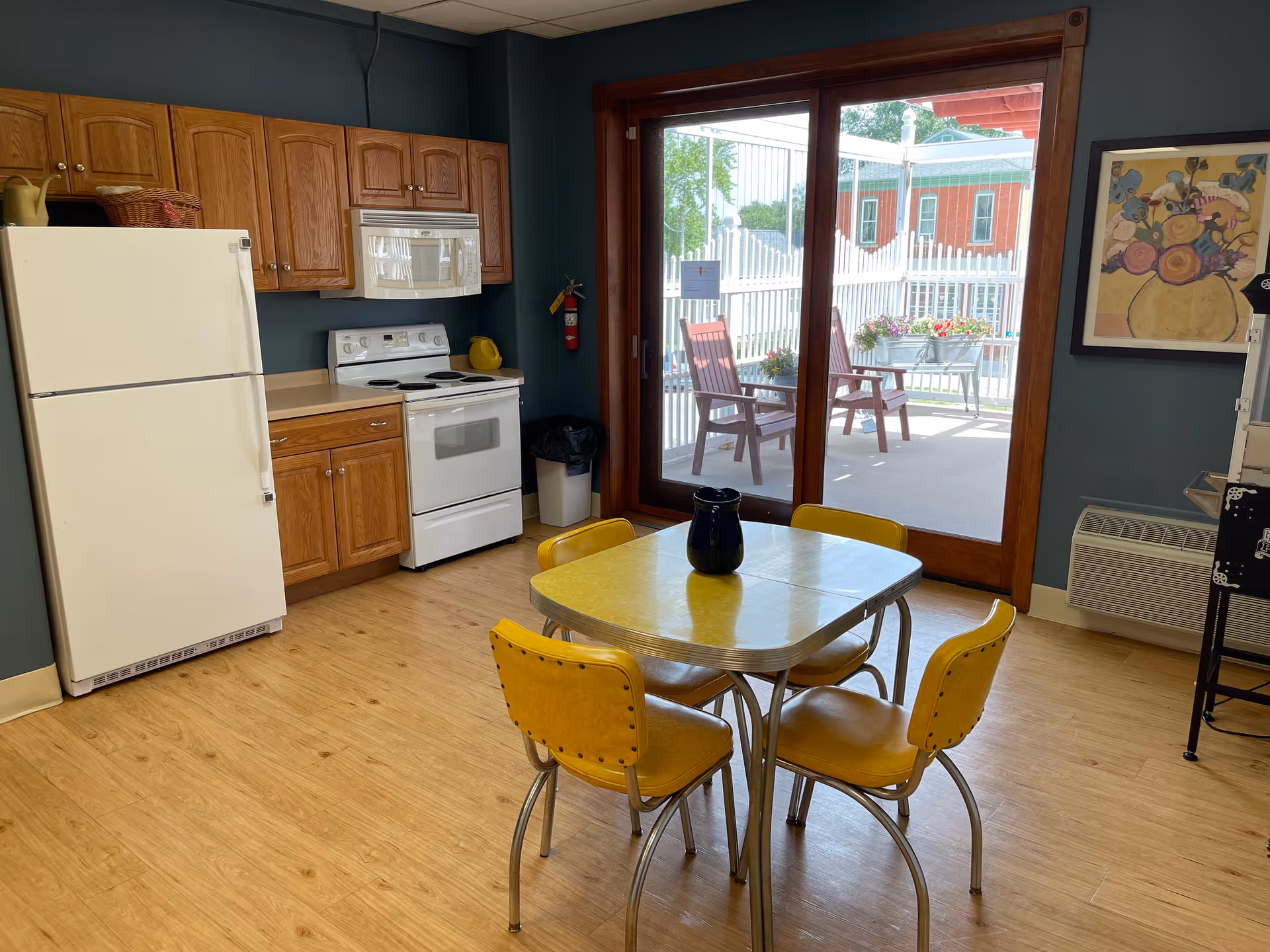 A kitchen area with wooden cabinets, a white refrigerator, stove, and microwave. In front of the kitchen is a small yellow dining table with four matching yellow chairs. A black pitcher is on the table. Large sliding glass doors lead to an outdoor patio with chairs and flower pots. A colorful framed painting hangs on the wall to the right.