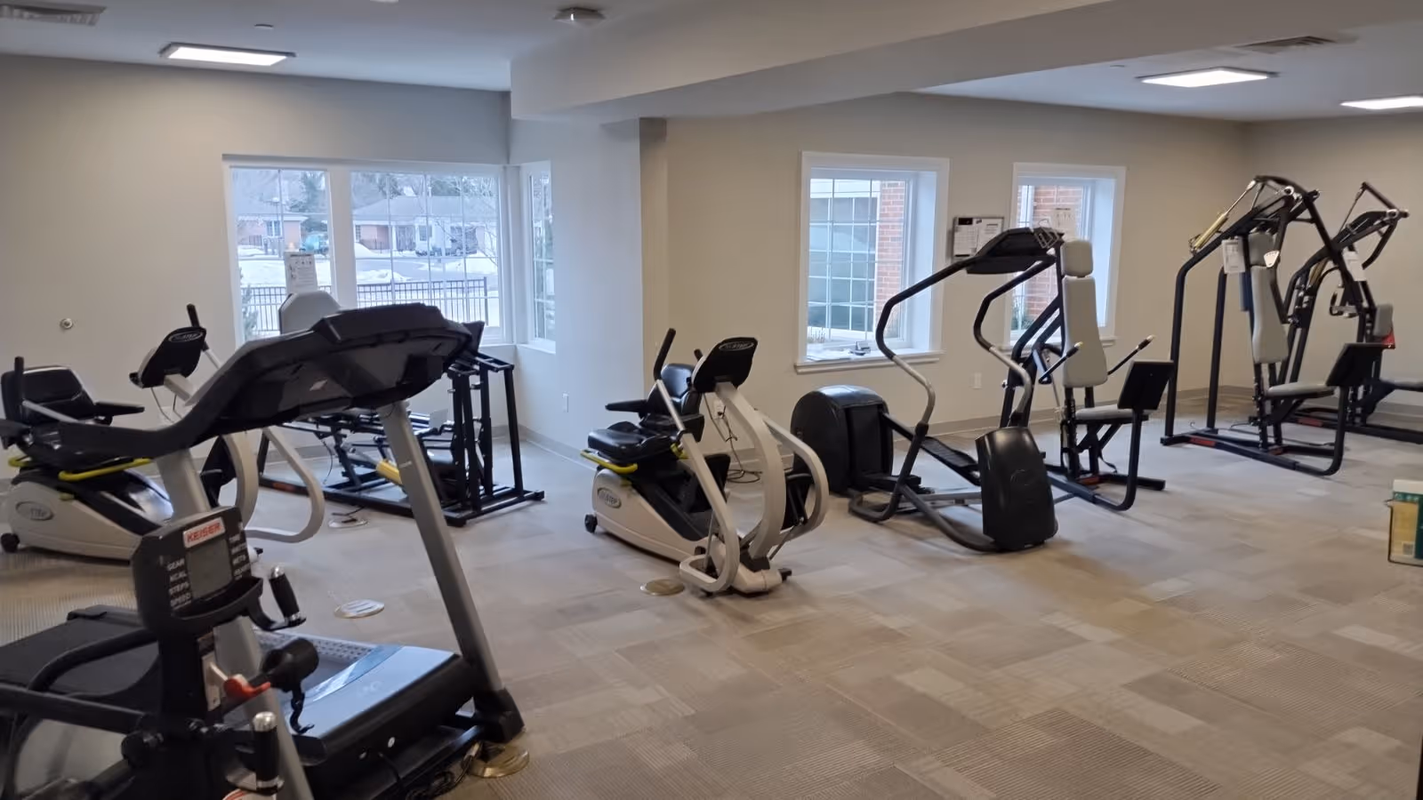 A fitness room with various exercise equipment including treadmills, stationary bikes, elliptical machines, and weight machines. The room has large windows letting in natural light and light-colored walls and carpeted flooring.