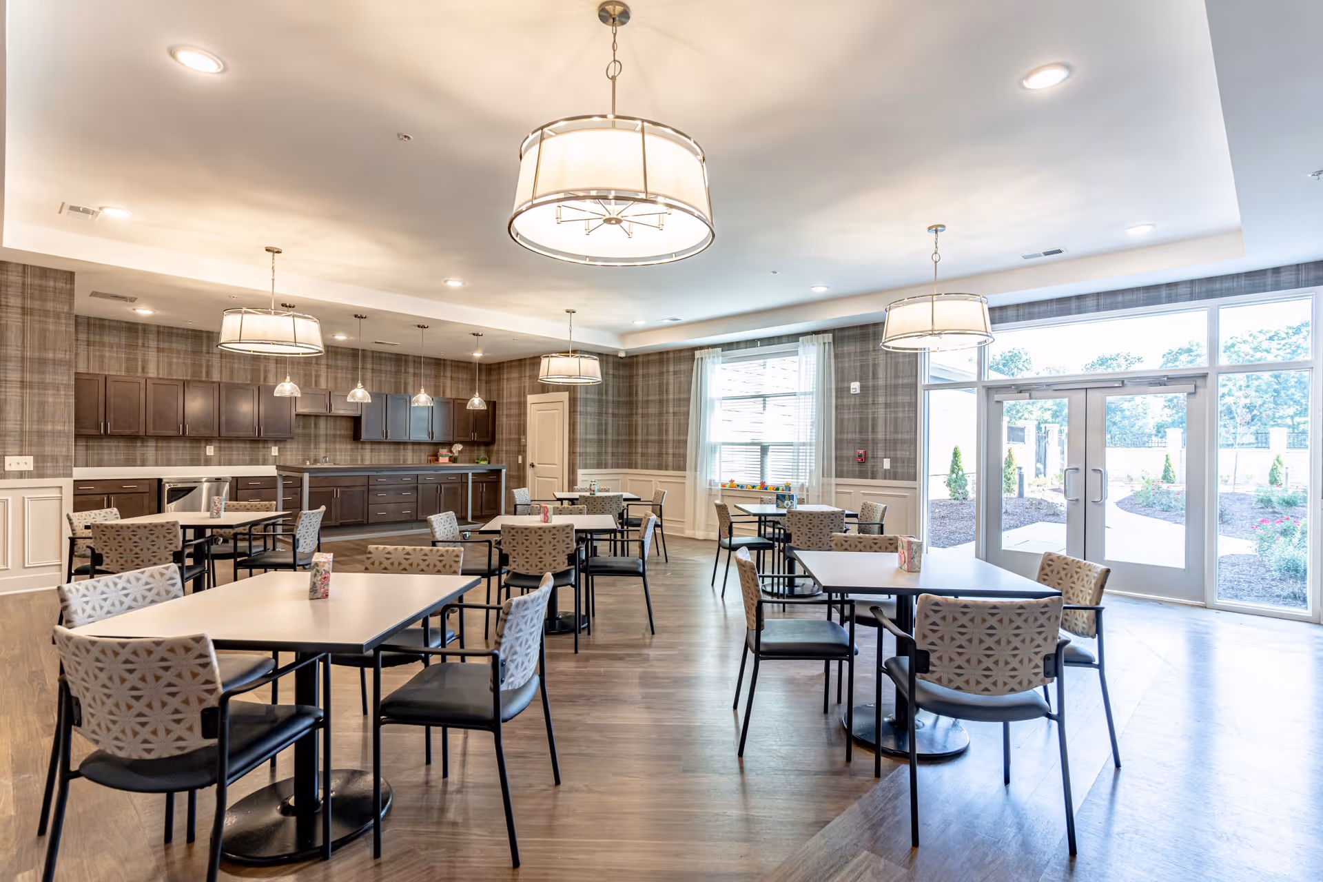 A bright and spacious dining room with multiple square tables and patterned chairs. The room features large windows and glass doors letting in natural light, modern pendant lights hanging from the ceiling, and a kitchen area with dark wood cabinets and stainless steel appliances along one wall.