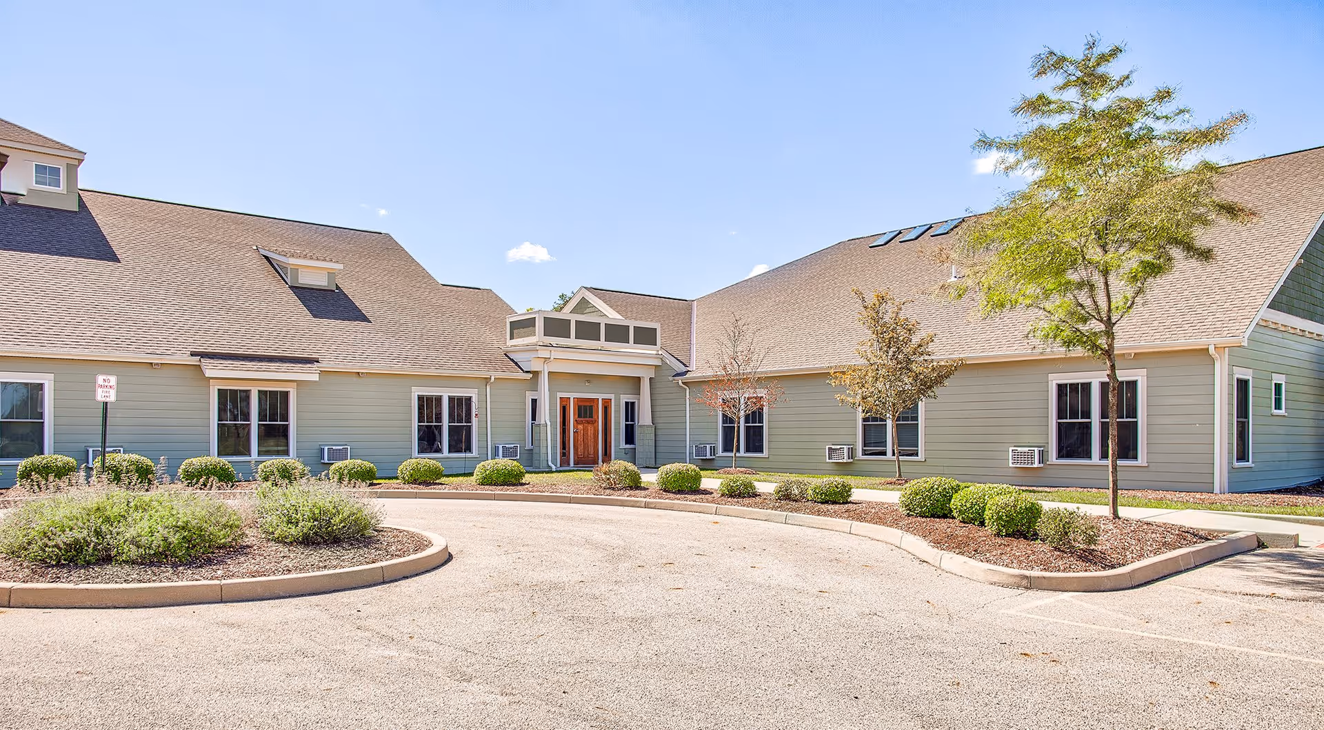 Front exterior of a single-story senior living building with a circular driveway, landscaped shrubs, and a covered entrance.