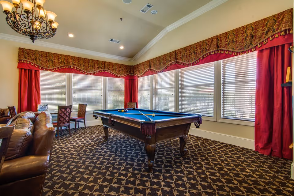 A well-lit recreational room with a pool table in the center, surrounded by large windows with red curtains and patterned valances. There are several chairs and a leather sofa along the left side, and a decorative chandelier hangs from the ceiling.