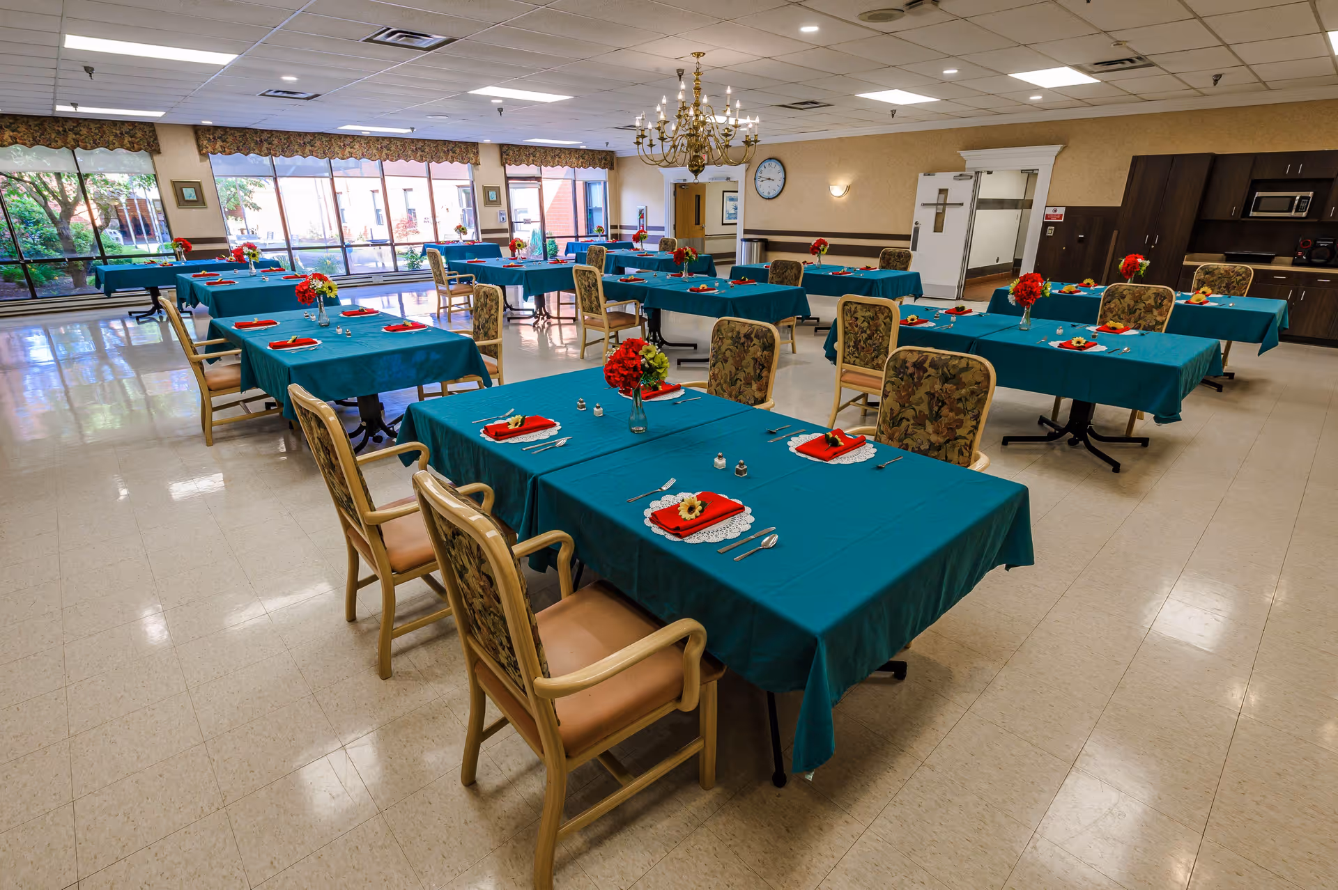 A spacious dining room with multiple tables covered in teal tablecloths, each set with red napkins, silverware, and small floral centerpieces. The room has large windows letting in natural light, floral-patterned chairs, a chandelier, and a clock on the wall.