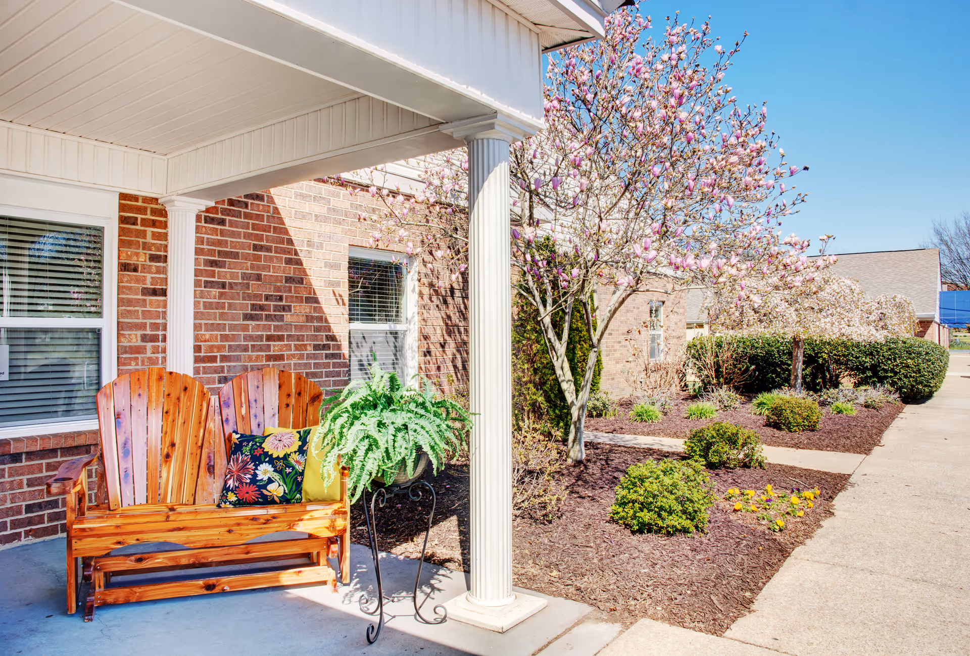 Outdoor seating area at Red Cedar Glen featuring a wooden bench with colorful cushions under a covered porch. Nearby is a potted fern on a decorative metal stand. The scene includes a brick building wall, blooming trees with pink flowers, neatly trimmed bushes, and a clear blue sky.