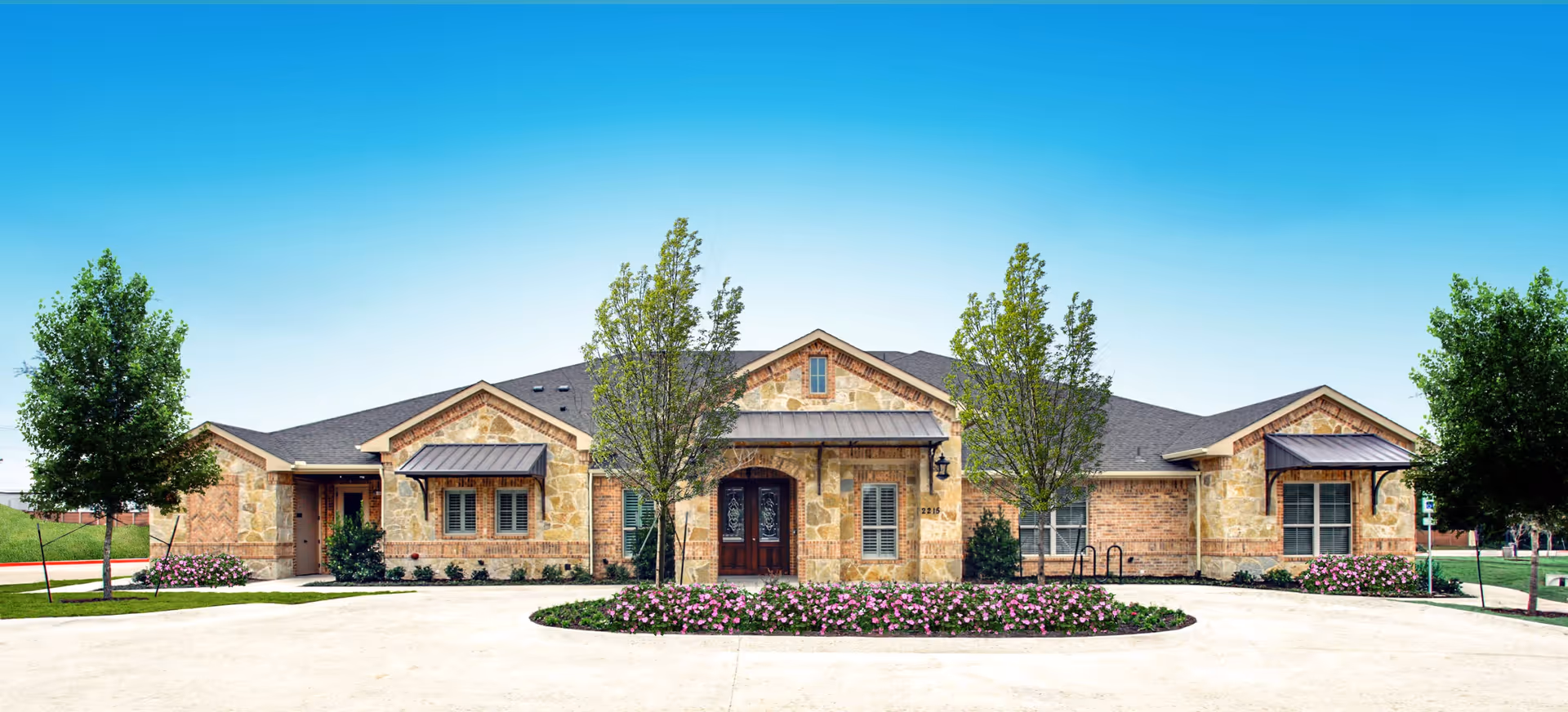 Front exterior view of a single-story stone and brick building with a dark shingled roof, three trees, and flower beds in front under a clear blue sky.