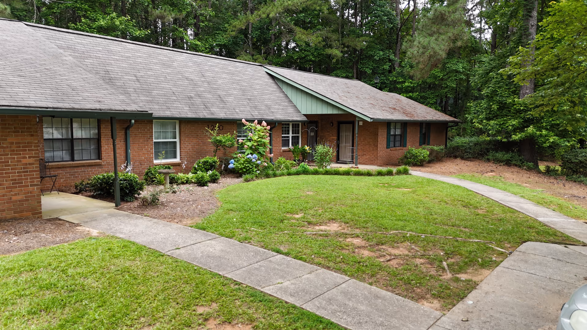 Exterior view of a single-story brick building with a gray shingled roof, surrounded by green grass, shrubs, and trees. A concrete walkway curves in front of the building, leading to the entrance doors. The area appears to be peaceful and wooded.