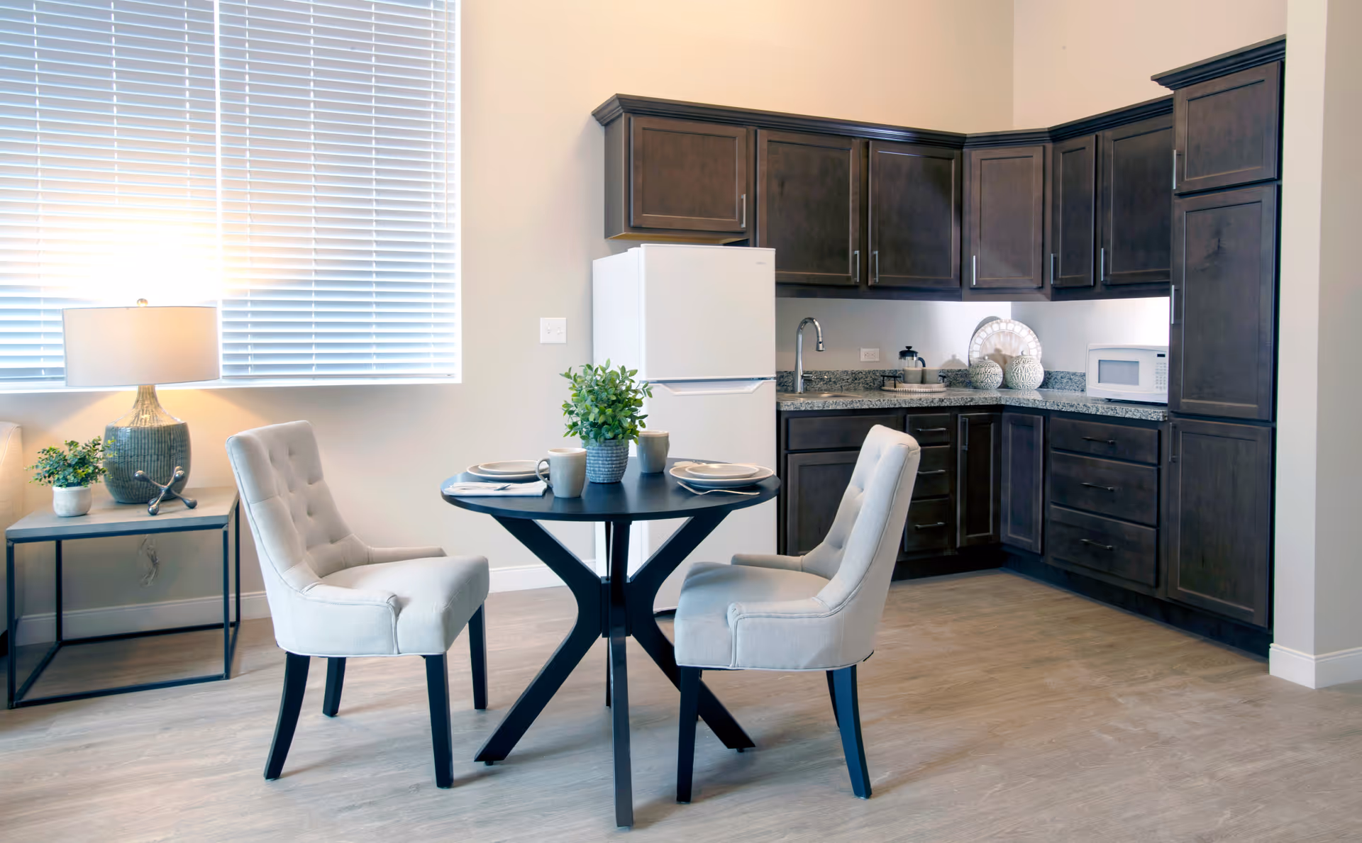 Bright kitchenette with dark wood cabinets, a small round dining table set with two upholstered chairs, a refrigerator, and a lamp on a side table.