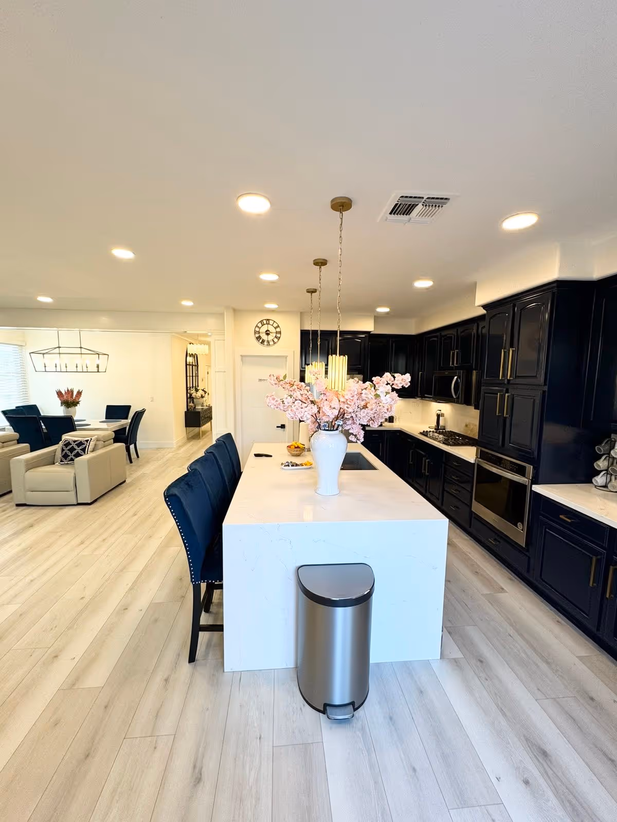 Modern kitchen with a large white island countertop featuring a vase with pink flowers, surrounded by blue cushioned chairs. The kitchen has dark cabinetry with gold handles, built-in oven, microwave, and a gas stove. The floor is light wood, and the ceiling has multiple recessed lights. In the background, there is a dining area with a table and chairs, and a living area with beige armchairs.