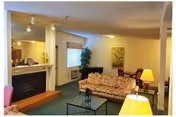 Interior view of a living room area in Leisure Manor Apartments featuring a floral patterned sofa, glass coffee table, two table lamps, a fireplace, a window with a valance, a potted plant, and a dining table with chairs in the background.