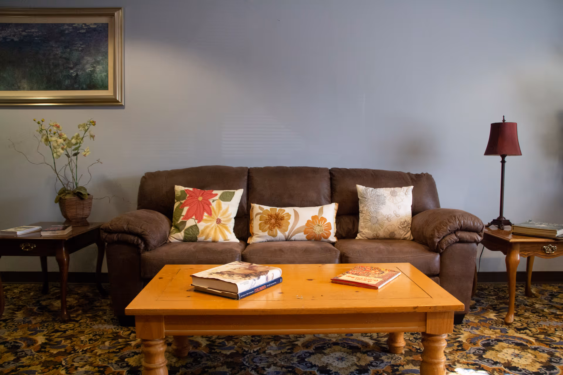 A cozy living room with a brown sofa adorned with three floral-patterned pillows. In front of the sofa is a wooden coffee table with two books on it. On either side of the sofa are wooden side tables, one with a potted plant and the other with a red lampshade. A framed painting hangs on the wall above the left side table, and the floor is covered with a patterned carpet.