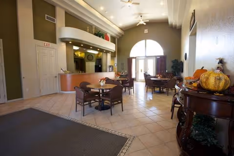 A spacious and well-lit common area with high ceilings, several round tables with chairs, a reception desk, and large arched windows letting in natural light. There is a decorative table with pumpkins and fall-themed decor on the right side.