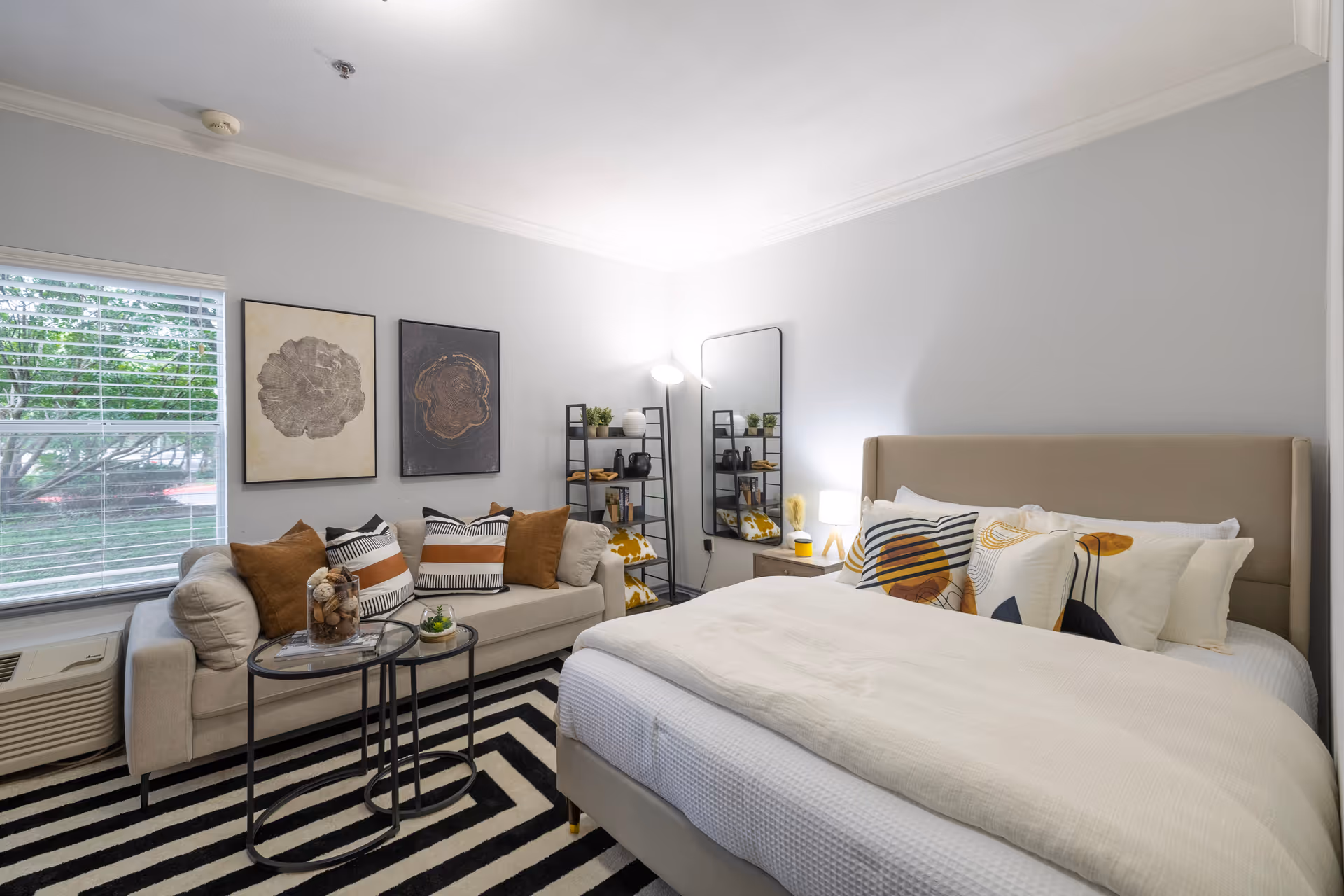 A cozy bedroom featuring a large bed with white and patterned pillows, a beige upholstered headboard, and a white comforter. Next to the bed is a small nightstand with a lamp. Across from the bed is a beige sofa adorned with multiple decorative pillows in brown, white, and striped patterns. Two round nesting coffee tables with decorative items sit in front of the sofa on a black and white striped rug. The room has light gray walls, two framed abstract art pieces above the sofa, a tall mirror, a black shelving unit with plants and decor, and a window with white blinds showing greenery outside.
