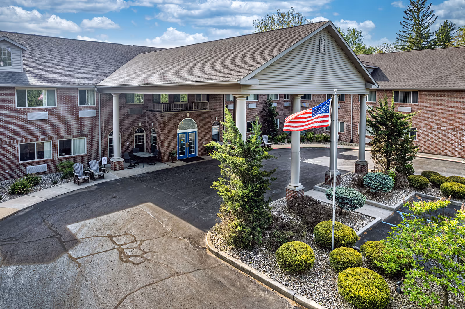 Front exterior view of a senior living facility with a covered entrance supported by white columns, an American flag on a flagpole, well-maintained landscaping with bushes and trees, and a brick building with multiple windows and blue double doors.