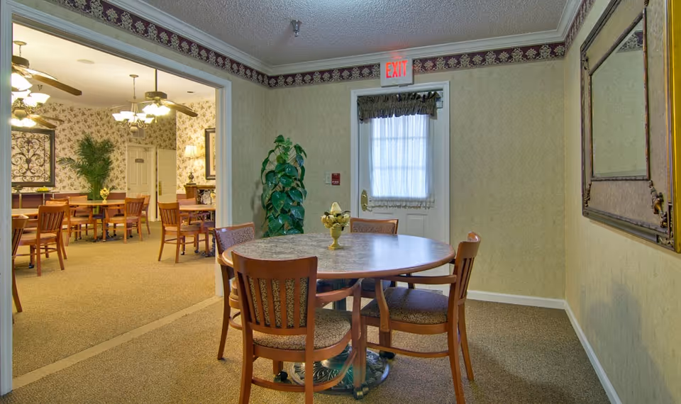 Interior view of a senior living facility dining area with a round table and four wooden chairs in the foreground. In the background, there is a larger dining room with multiple tables and chairs, ceiling fans with lights, and floral wallpaper. A potted plant is placed near a door with an exit sign above it. A large mirror hangs on the right wall.