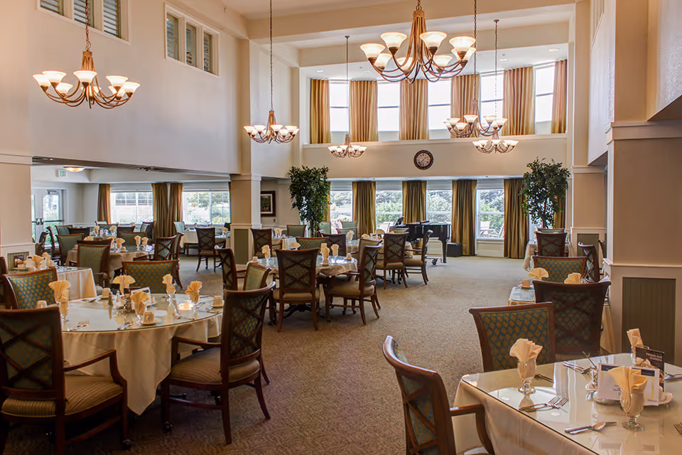 A spacious, well-lit dining room with multiple round tables covered with white tablecloths, each set with folded napkins, glassware, and silverware. The room features high ceilings with large windows draped with gold curtains, several chandeliers hanging from the ceiling, and green plants placed near the windows.