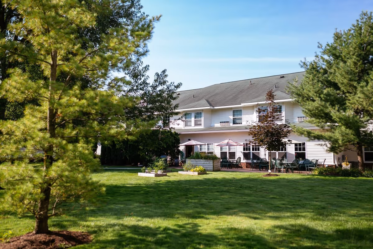 Outdoor view of Woodland Village facility showing a well-maintained grassy lawn with several trees and raised garden beds. The building in the background has white siding and multiple windows, with a patio area featuring tables, chairs, and umbrellas.