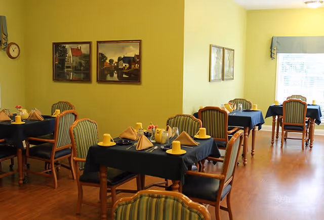 A dining room with several wooden tables covered with dark blue tablecloths, each set with yellow cups, folded napkins, silverware, and small flower arrangements. The room has light yellow walls, wooden flooring, framed paintings on the walls, and windows with blue valances letting in natural light.