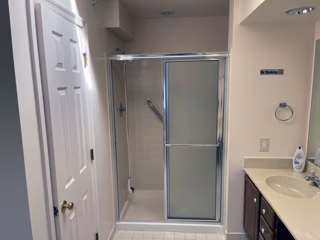 Bathroom with a shower enclosure featuring sliding frosted glass doors, a white tiled shower area with a grab bar, a sink with a beige countertop and dark wooden cabinets, a towel ring, a bottle of lotion on the counter, and a 'No Smoking' sign on the wall.
