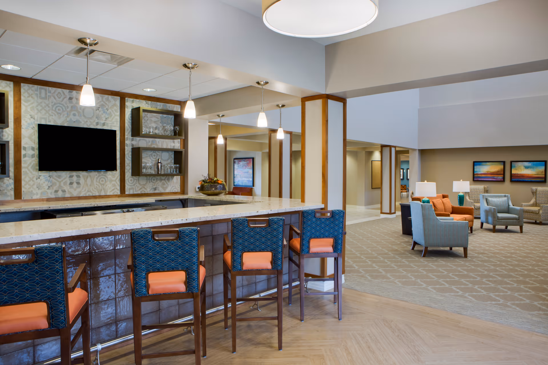 Bright communal lounge with a long bar and high stools in the foreground and seating clusters of armchairs and tables in the background.