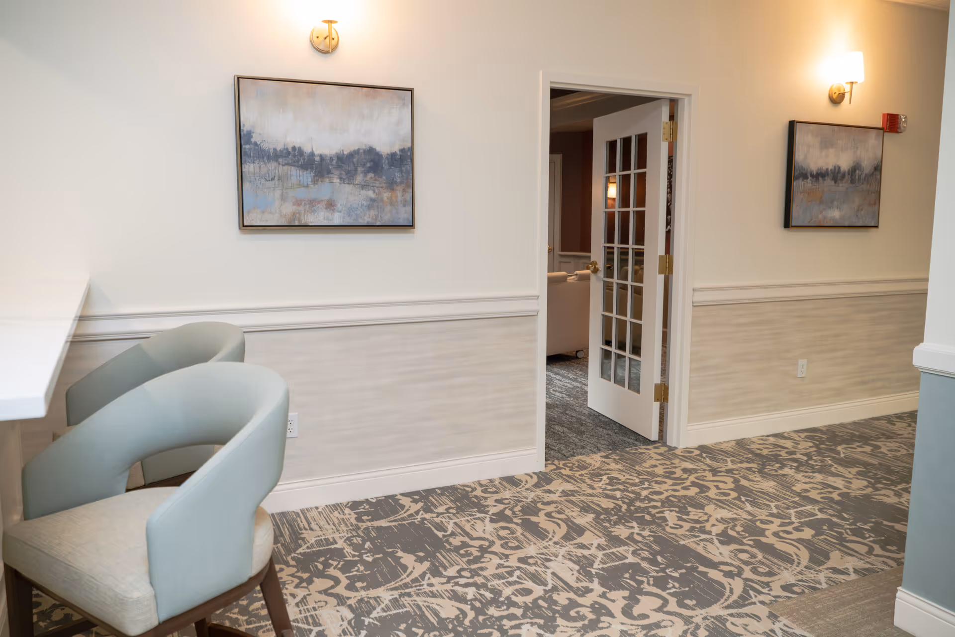 Interior hallway area with patterned carpet, two light blue cushioned chairs at a white counter on the left, and a white door with glass panes open to another room. Two abstract paintings hang on the walls, which are painted white with a beige wainscoting. Wall sconces provide lighting.