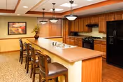 Communal kitchen and dining area with a long island lined with four bar stools, wood cabinetry, black appliances, and pendant lighting.