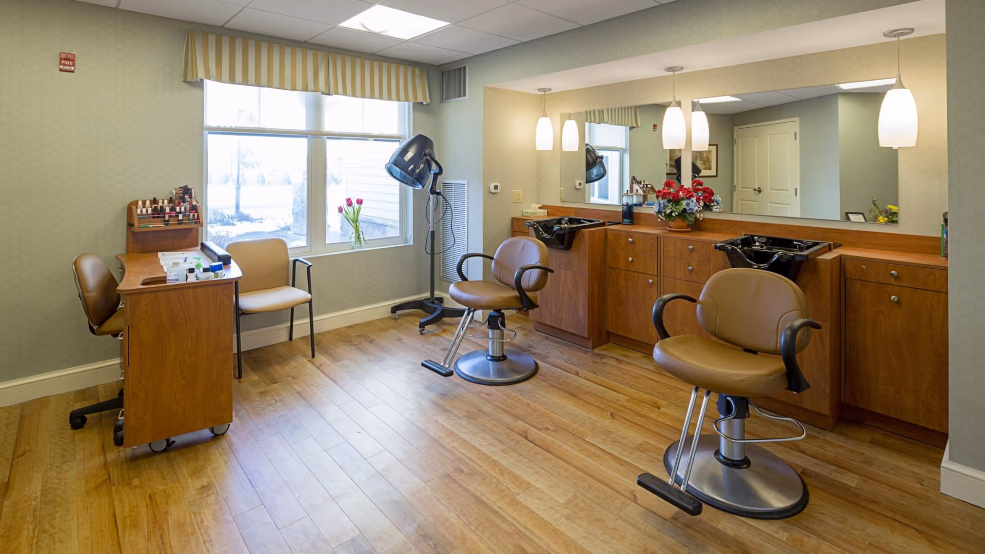 Interior view of a salon area in a senior living facility with two brown salon chairs in front of a large mirror and two black hair washing sinks. There is a wooden manicure table with nail polish and supplies near a window with striped yellow and white valance. The room has wooden flooring and soft lighting from pendant lamps.