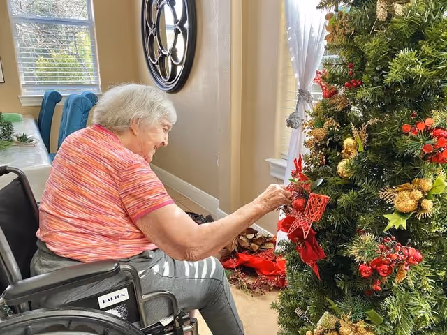 An elderly woman in a wheelchair decorates a Christmas tree with red and gold ornaments in a well-lit room with beige walls and a large window with white curtains.