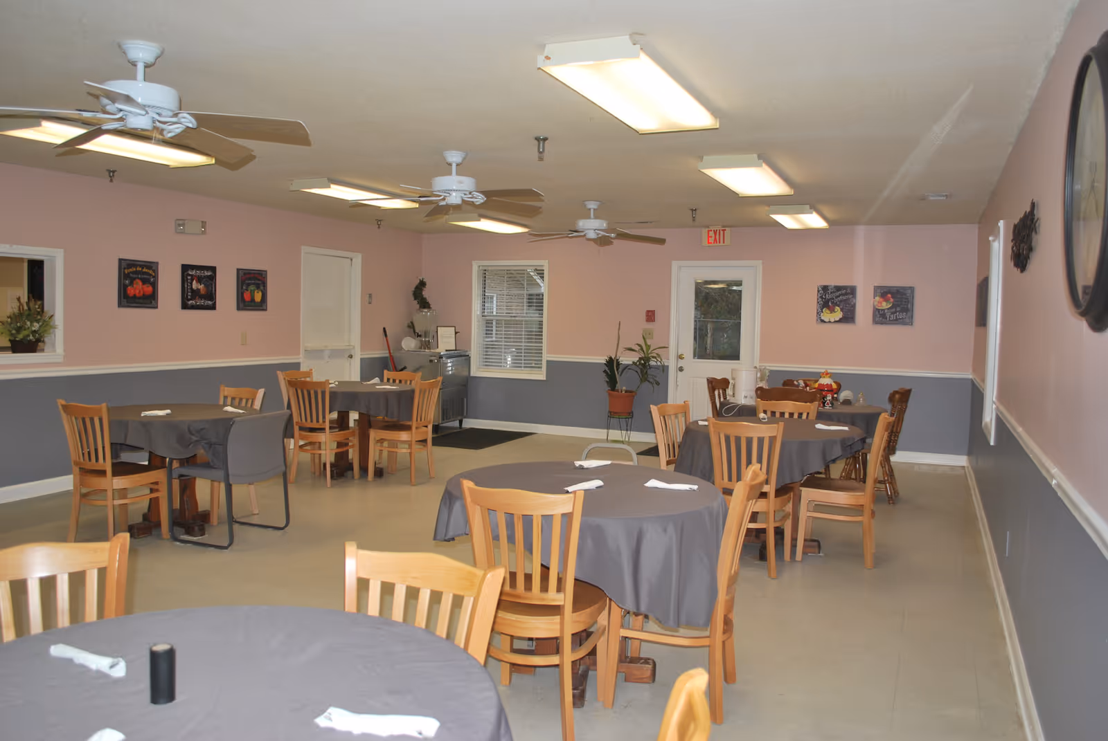 A dining room with several round tables covered with gray tablecloths and wooden chairs around them. The walls are painted pink on the upper half and gray on the lower half, decorated with framed pictures. Ceiling fans and fluorescent lights are mounted on the ceiling. There is a door and a window on the far wall, along with some potted plants and a small cabinet.