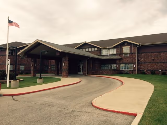 Front exterior view of Canoe Brook Assisted Living-Catoosa building with a covered entrance, brick facade, multiple windows, an American flag on a flagpole, and a curved driveway with red-painted curbs.