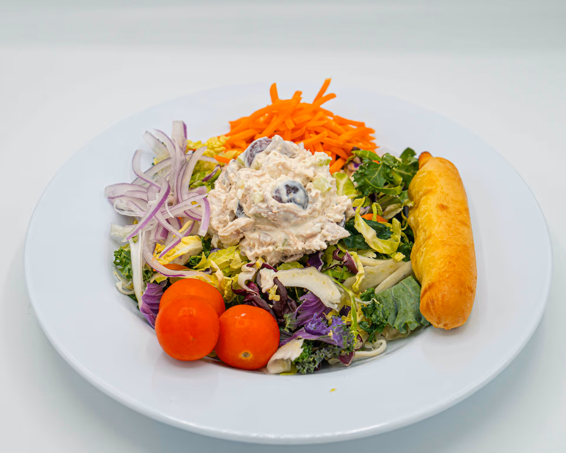 A white plate with a fresh salad consisting of leafy greens, shredded carrots, sliced red onions, two cherry tomatoes, and a scoop of chicken salad with grapes, accompanied by a breadstick on the side.