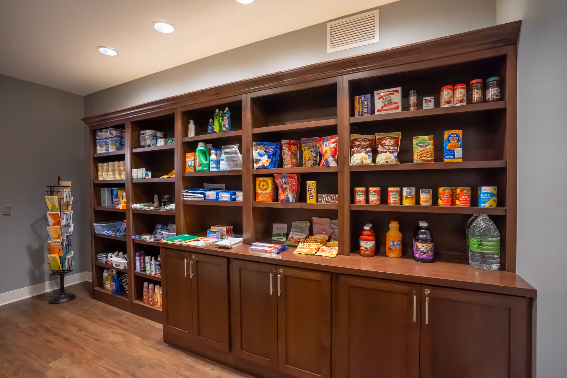 A large wooden shelving unit filled with various grocery and household items including canned goods, snacks, cleaning supplies, and bottled drinks in a well-lit room with wood flooring and gray walls.