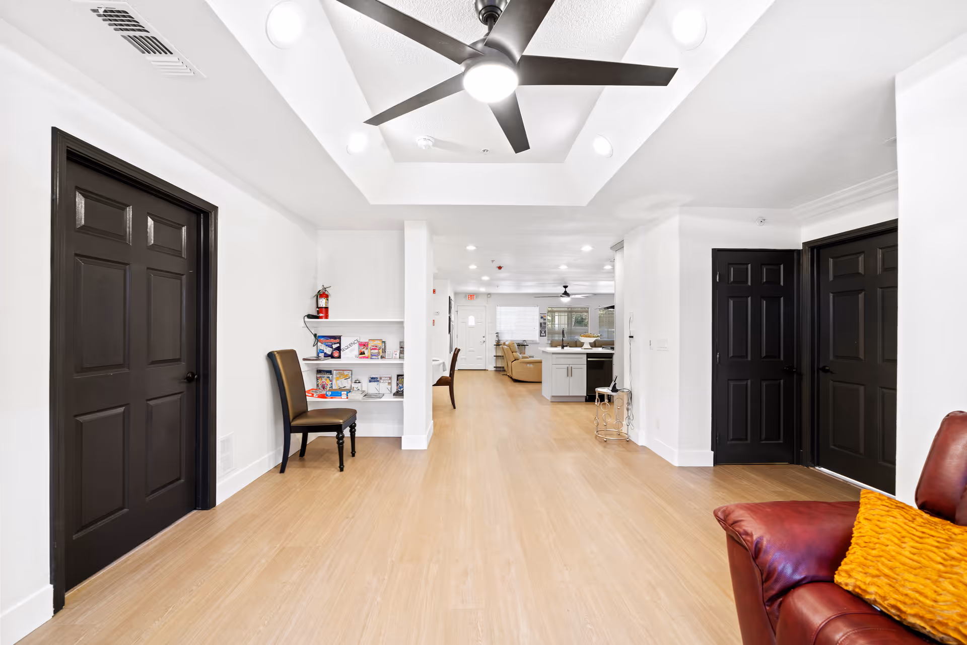 Interior view of a senior living facility hallway with light wood flooring, white walls, and black doors. A ceiling fan with lights is mounted on a recessed ceiling. There is a brown chair next to a small built-in shelf holding brochures and a fire extinguisher. In the background, a living area with beige sofas and a kitchen island is visible. A red leather chair with a yellow cushion is partially visible in the foreground on the right.
