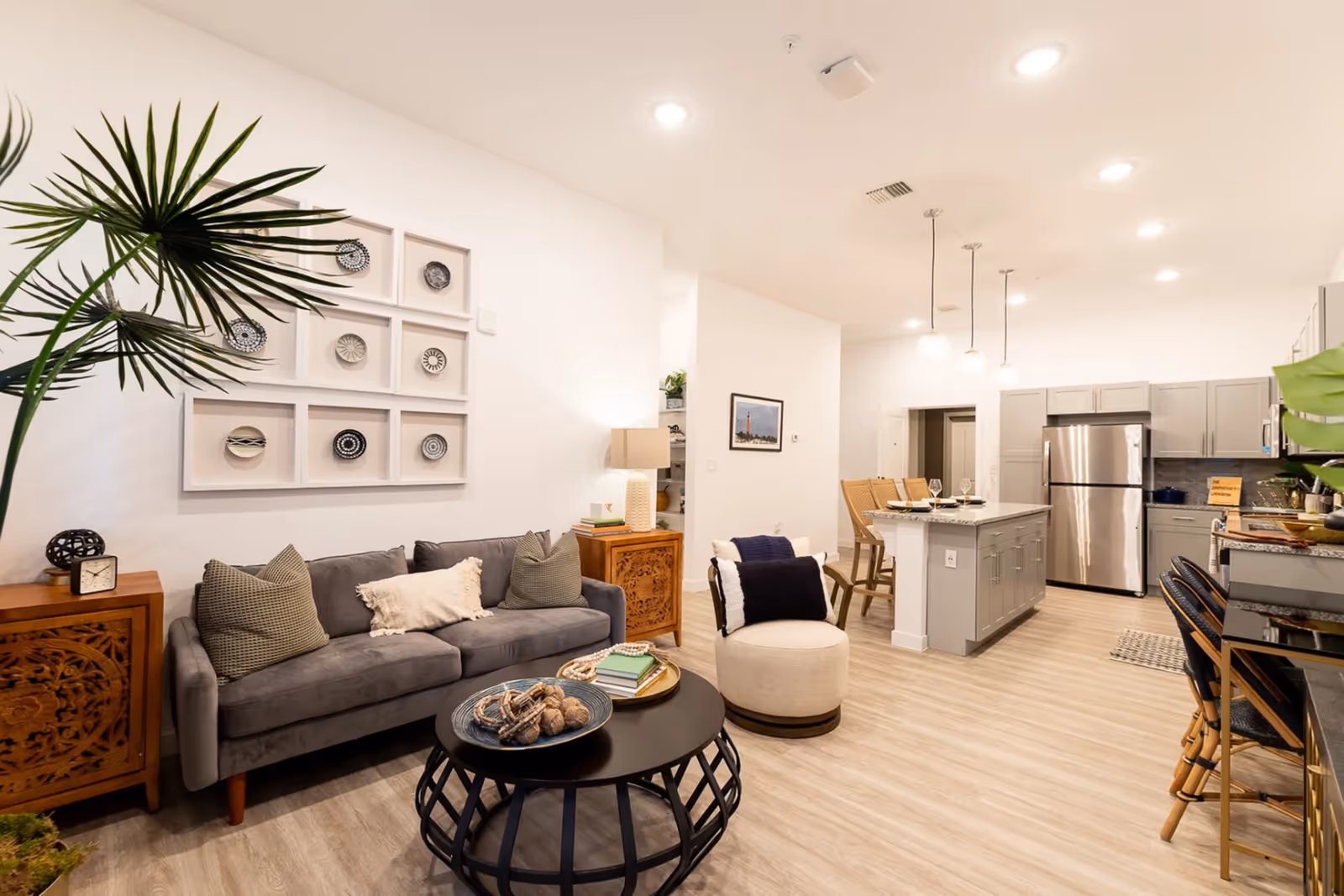 Open-plan living area with a gray sofa, round black coffee table, decorative wall art, and a kitchen island with stainless refrigerator in the background.