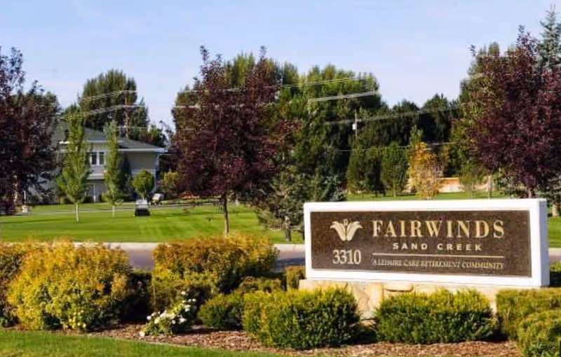 Stone entrance sign reading "Fairwinds Sand Creek" surrounded by landscaped shrubs and trees with a building in the background.