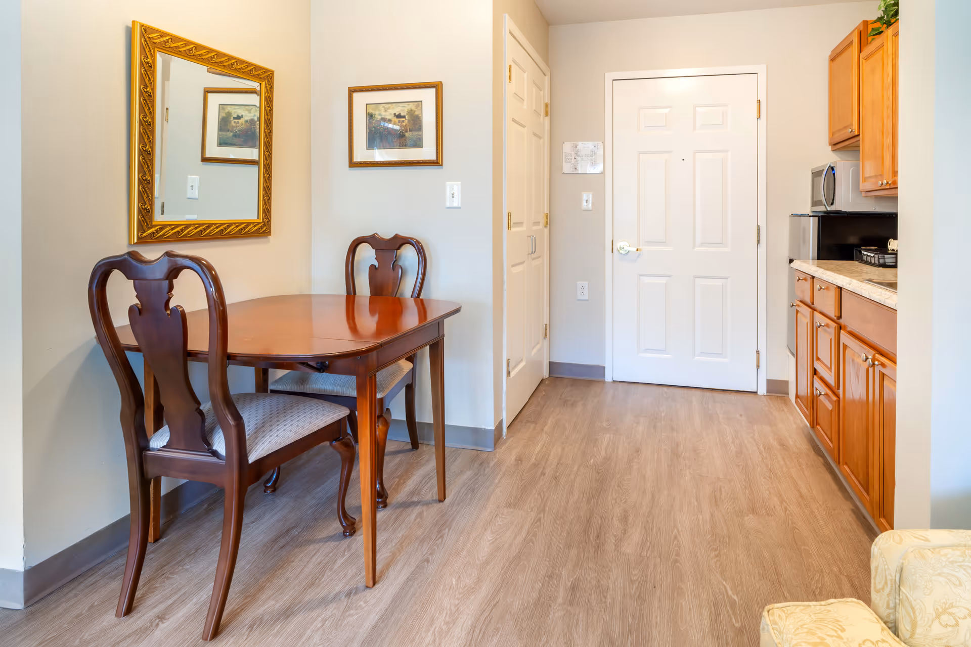 A small dining area with a wooden table and two matching chairs with cushioned seats. A large ornate gold-framed mirror and a framed picture hang on the wall above the table. To the right, there is a kitchenette with wooden cabinets, a microwave, and a countertop. A white door and a closet with double doors are visible in the background. The floor is covered with light wood laminate.