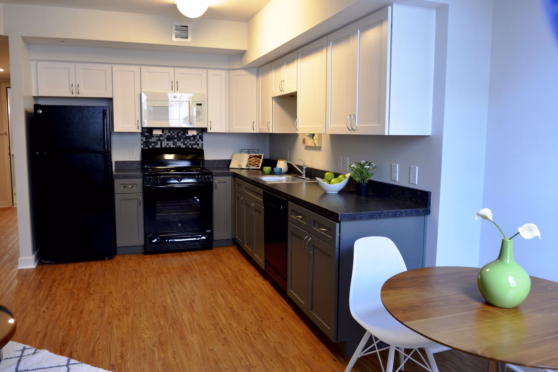 Modern kitchen with black refrigerator, black stove, white microwave, and gray and white cabinets. The kitchen has a black countertop with a sink, a bowl of green apples, and a small potted plant. There is a round wooden table with a white chair and a green vase with white flowers on it. The floor is wood with a light brown finish.