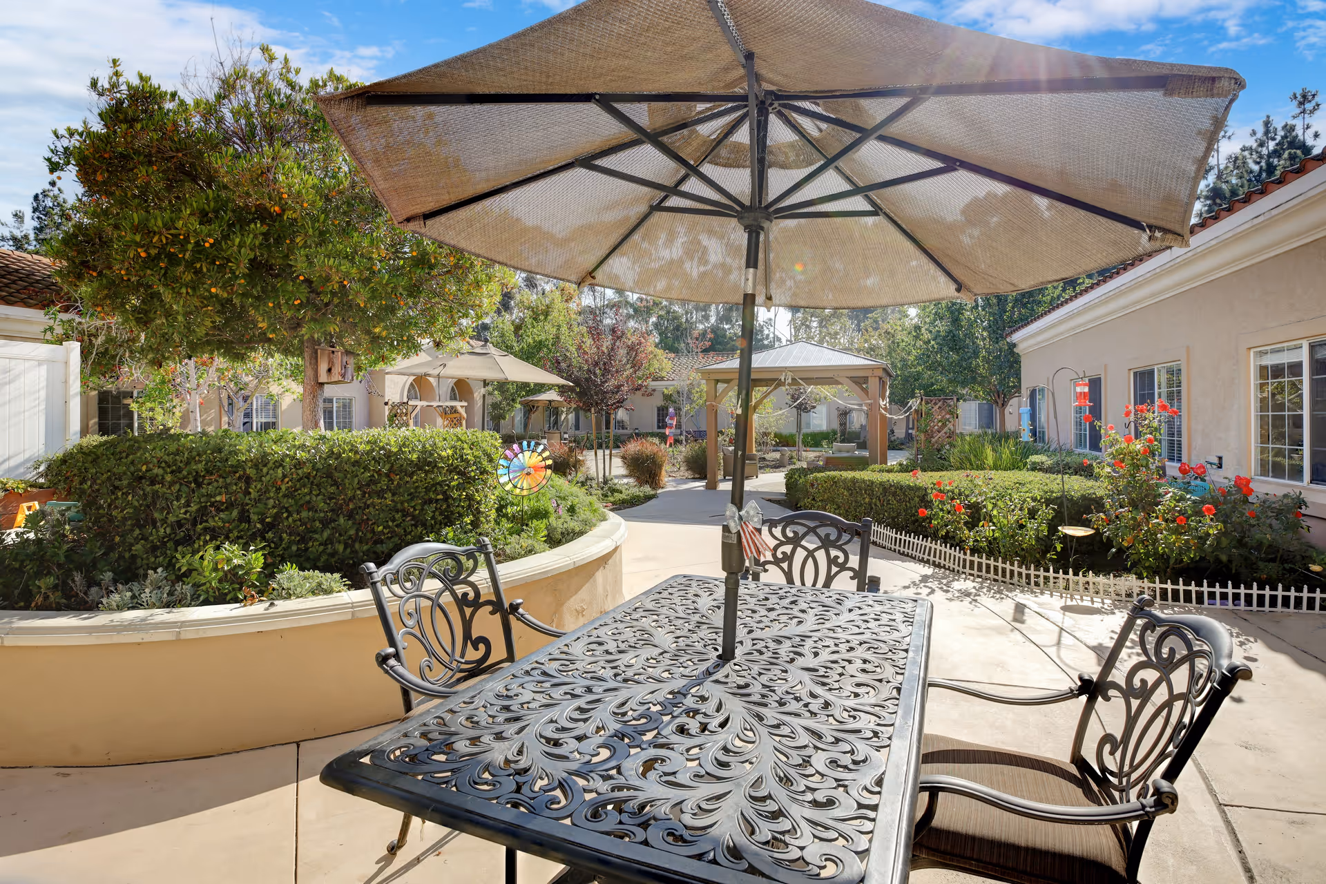 Outdoor patio area with a decorative metal table and chairs under a large beige umbrella. Surrounding the patio are well-maintained bushes, flowering plants, and trees. In the background, there are buildings with windows and a gazebo structure, all under a clear blue sky.