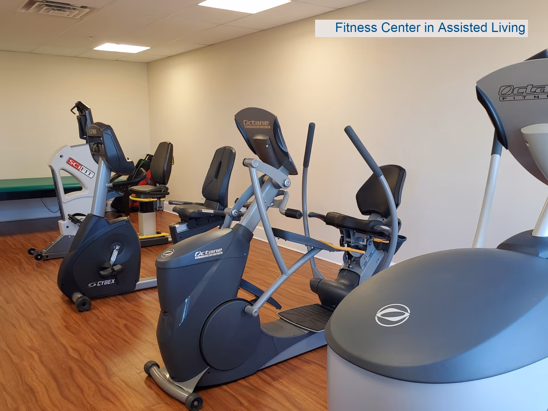 A fitness center room in an assisted living facility with several exercise machines including recumbent bikes and elliptical trainers on a wooden floor with plain white walls.