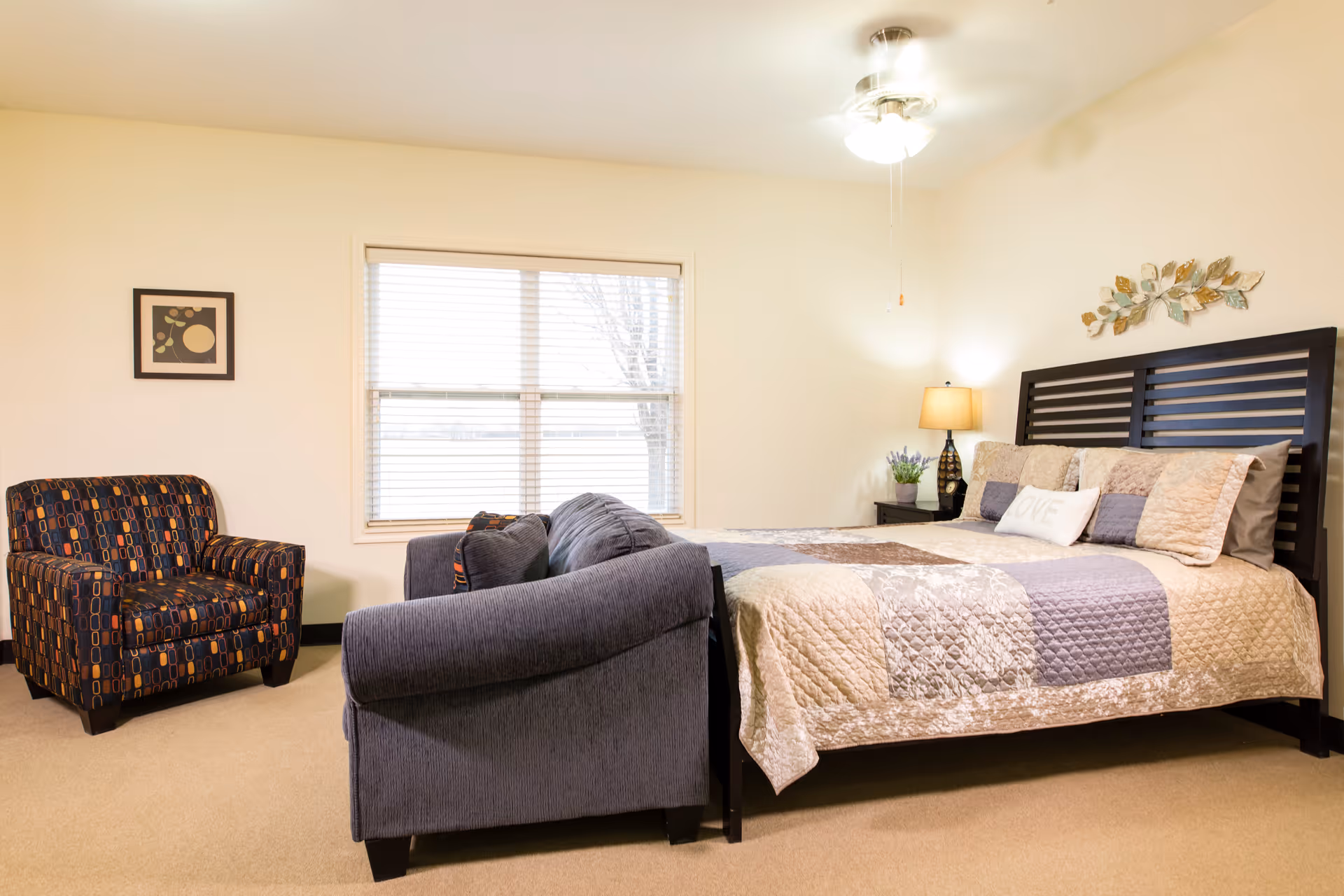 Well-lit bedroom featuring a bed with patterned quilt, a nightstand and lamp, a sofa and a patterned armchair beside a window.
