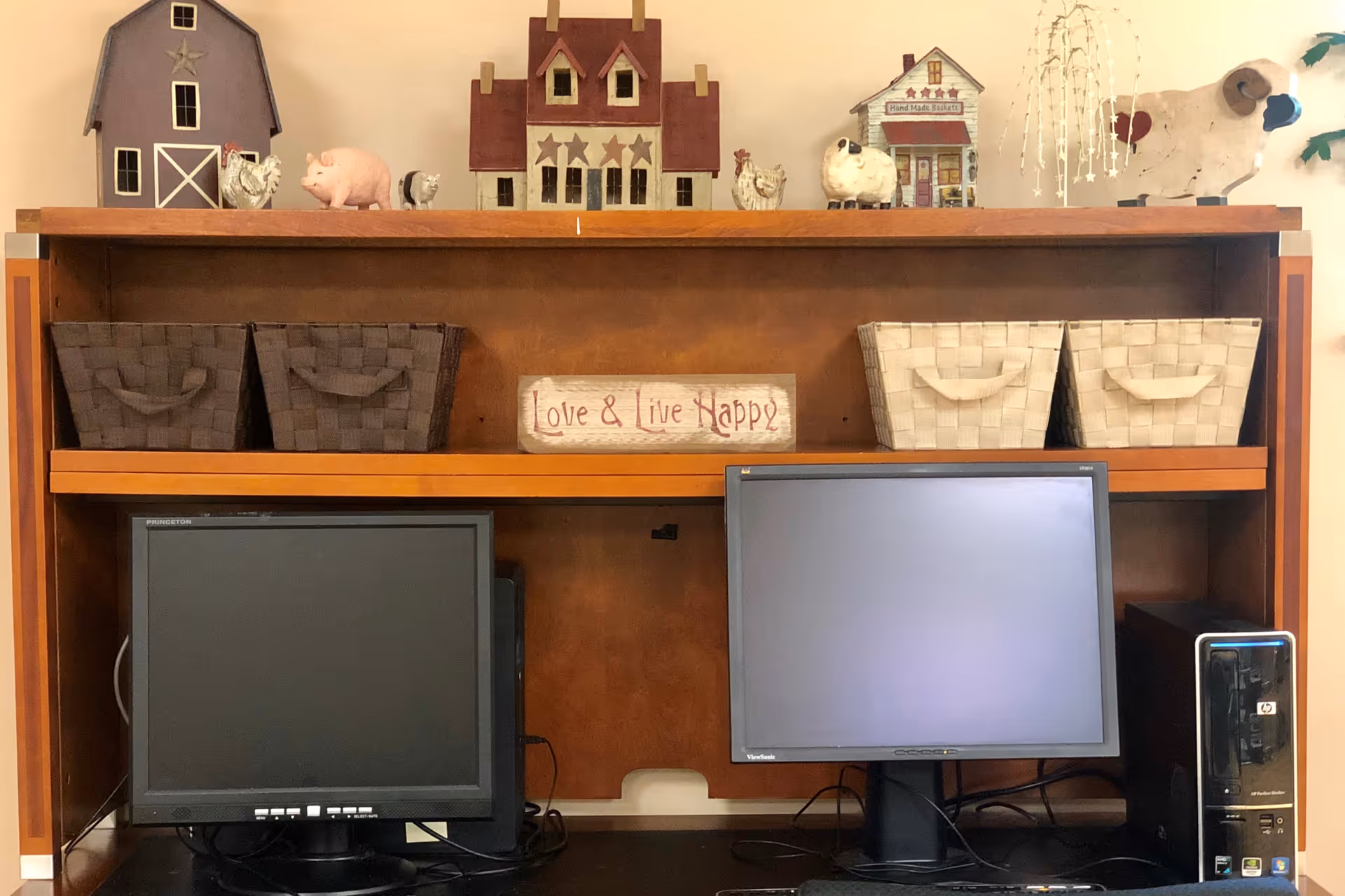 A wooden desk with two computer monitors and a desktop tower. Above the desk are four woven storage baskets and a decorative sign that reads 'Love & Live Happy'. On top of the shelf are various small decorative items including miniature houses, farm animals, and a hanging ornament.