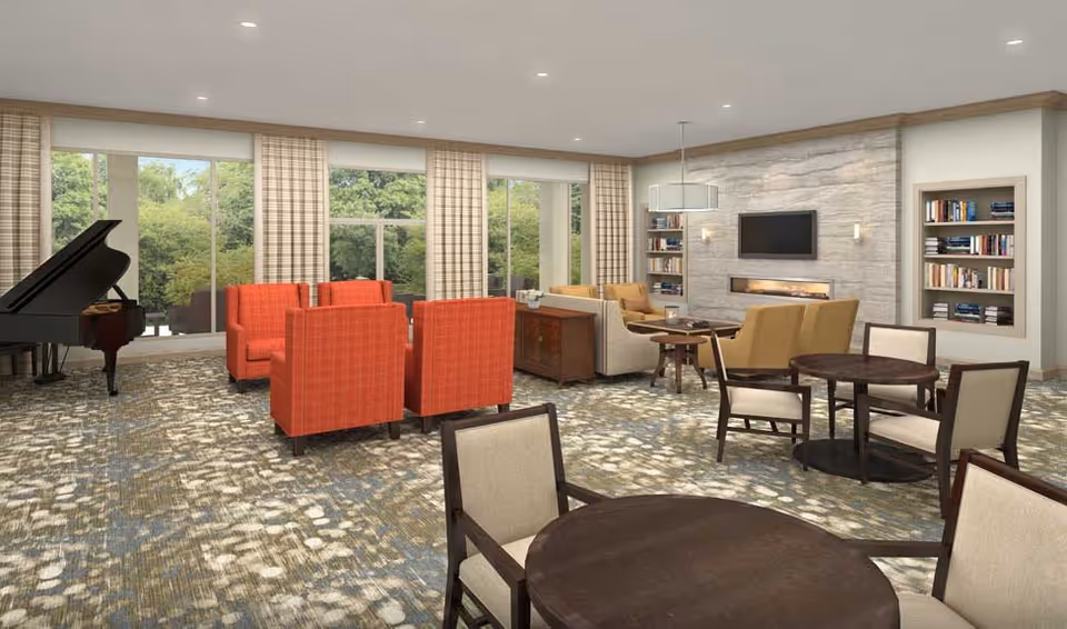 A spacious and well-lit living room area in a senior living facility featuring large windows with views of greenery outside. The room includes multiple seating arrangements with red and beige armchairs, round wooden tables with chairs, a grand piano in the corner, built-in bookshelves, a wall-mounted TV above a modern fireplace, and soft, patterned carpeting.